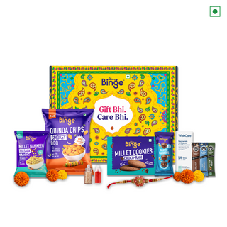A Binge brand gift basket set featuring a colorful patterned box, a packet of smoky BBQ quinoa chips, a box of Choco Ragi Millet cookies, a bottle of milk, and several packaged snack items arranged on a white background with decorative marigold flowers and a red and gold Rakhi.
