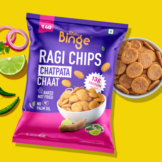 A bag of The Healthy Binge Ragi Chips Chatpata Chaat on a bright yellow background, with a bowl of round golden-brown chips and garnish ingredients like lime, onion, and green chilies nearby.