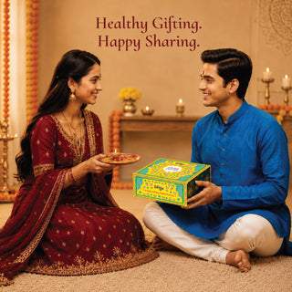 A man in a blue kurta and white pants sits cross-legged on a carpeted floor, holding a colorful decorative gift box, while a woman in a deep red embroidered traditional outfit kneels beside him holding a small plate. The background shows a warm, decorated indoor setting with candles and marigold garlands.
