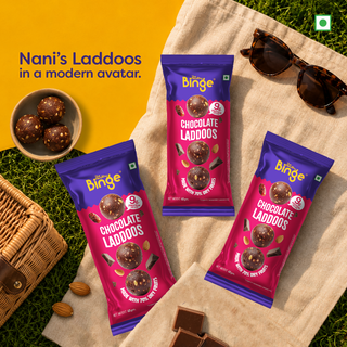Three packs of Nani's Laddoos by THB chocolate laddoos are arranged on a beige blanket with a yellow background, alongside sunglasses, a bowl of laddoos, almonds, and a wicker basket.