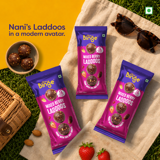 Three packs of Nani's Laddoos by THB are arranged on a beige blanket over green grass, with a wicker basket, strawberries, almonds, sunglasses, and a bowl of laddoos nearby. The background is split between yellow and green, with promotional text on the left.
