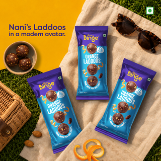 Three bags of Nani's Laddoo's by The Healthy Binge, orange-flavored is arranged on a beige picnic blanket with sunglasses, a small bowl of laddos, almonds, orange peel, and a wicker basket on a grassy surface.