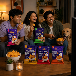 Three people sitting together in a cozy living room setting, enjoying Binge brand chips. Two of them are holding bags of chips (Ragi Chips, Miller Chips, Jowar Chips, and Quinoa Chips) while a bowl of chips and a lit candle sit on the wooden table in front of them.